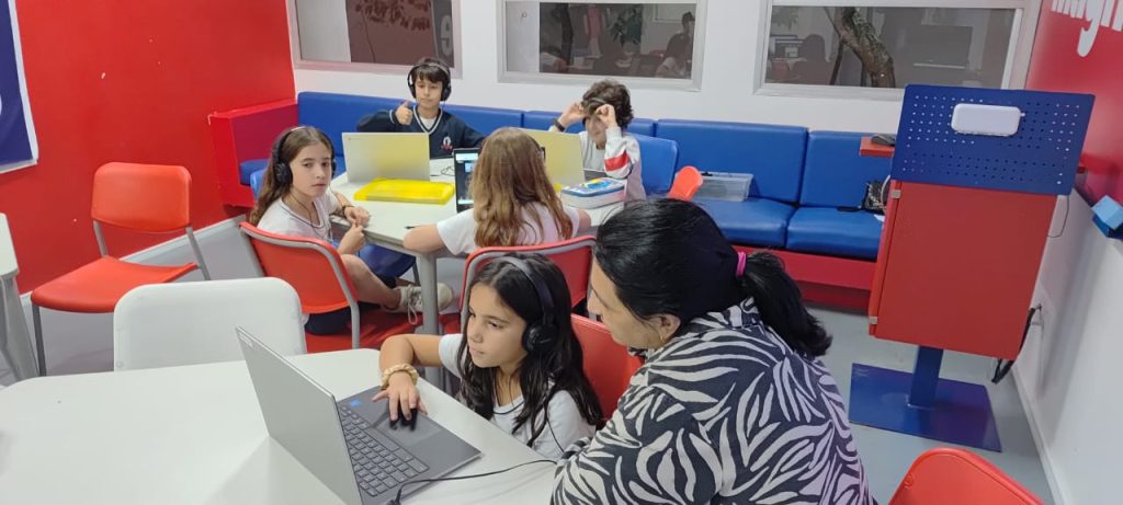 Student listening to a laptop while a teacher watches. Other students using laptops in the background.